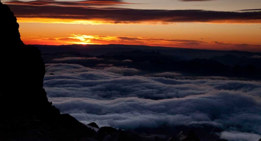 High above clouds, the sun rises or sets, causing the sky to appear in shades of orange and yellow.